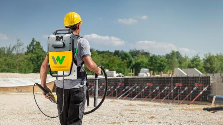 Construction worker carrying battery-powered Wacker Neuson high-frequency internal vibrator on a construction site.