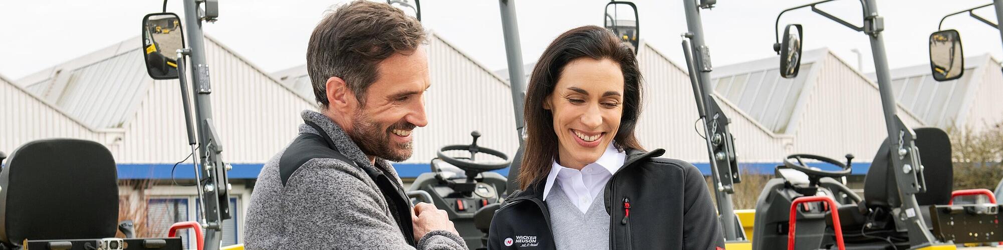 Une collaboratrice Wacker Neuson souriante et portant une tablette conseille un client souriant devant des machines de construction Wacker Neuson.