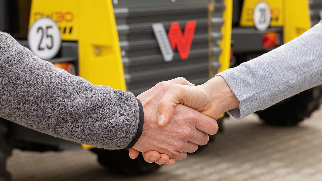 Podanie ruky medzi zamestnankyňou Wacker Neuson a zákazníkom, stavebné stroje Wacker Neuson v pozadí.