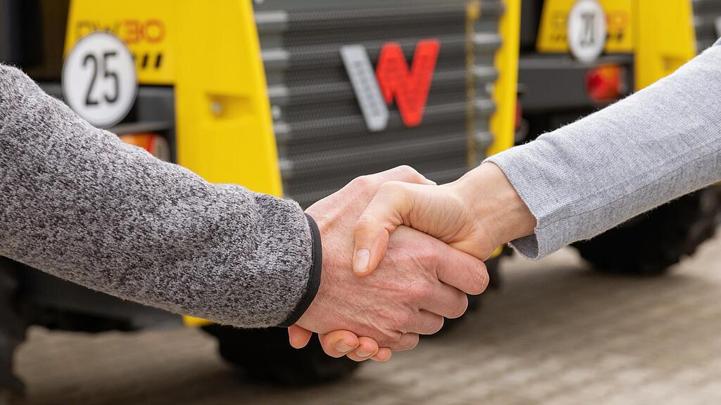 Handschlag zwischen Wacker Neuson Mitarbeiterin und Kunde, Wacker Neuson Baumaschinen im Hintergrund.