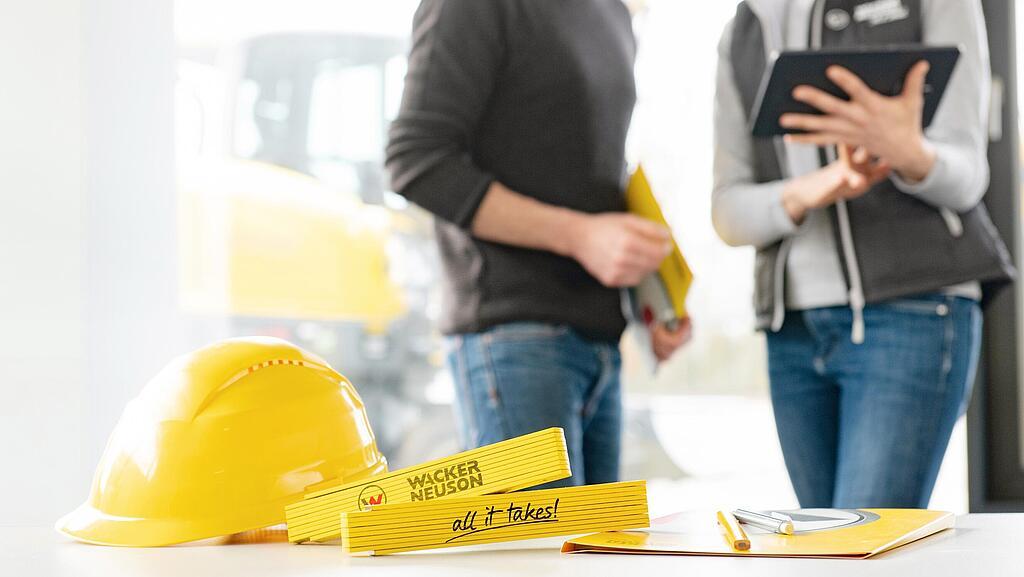 Wacker Neuson Merchandise Artikel liegen auf Tisch im Vordergrund, Wacker Neuson Mitarbeiter im Hintergrund.