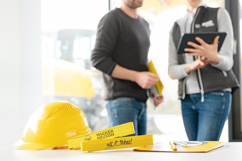 Wacker Neuson salgsartikkel ligger på bordet i forgrunnen, Wacker Neuson medarbeider i bakgrunnen.