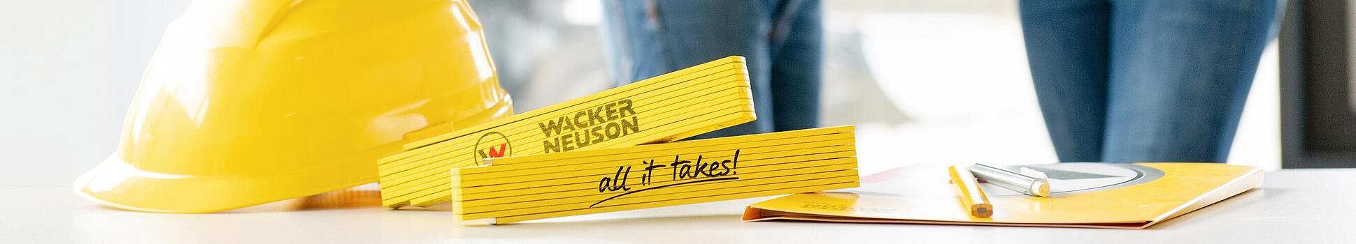 Wacker Neuson merchandise items lying on table in foreground, Wacker Neuson employees in background.