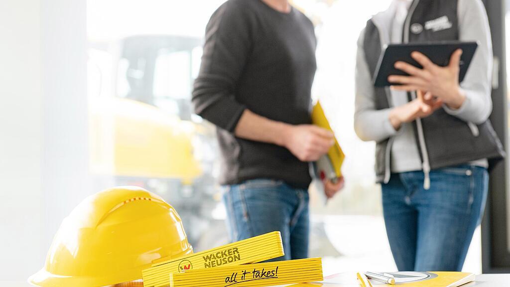 Des articles de merchandising Wacker Neuson sont posés sur une table au premier plan, collaborateur Wacker Neuson en arrière-plan.