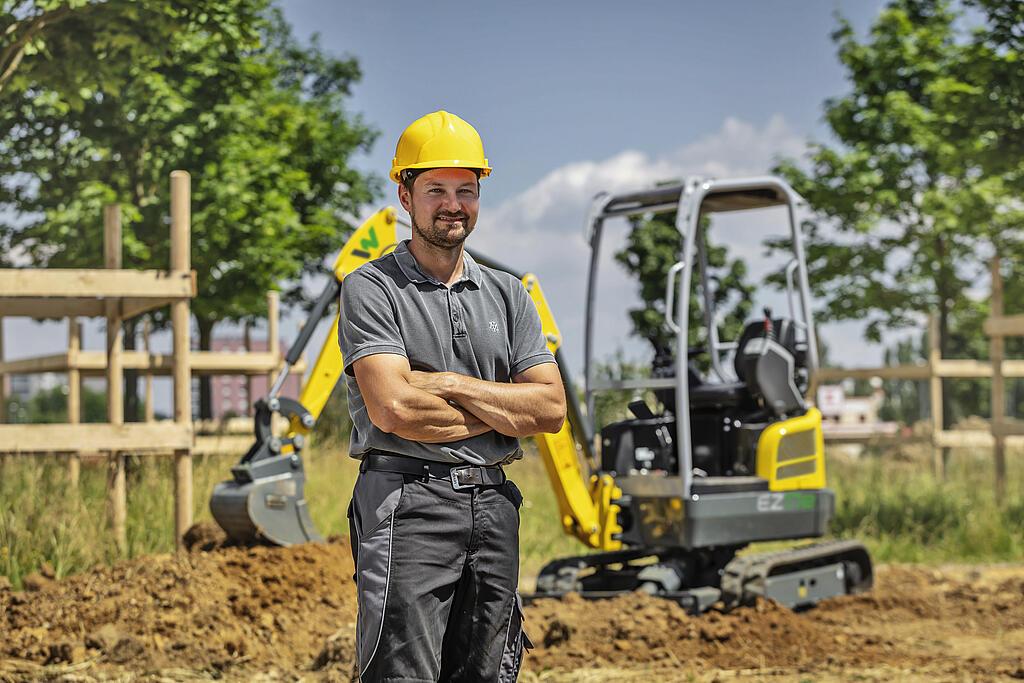 A man wearing a yellow hard hat stands in front of a Wacker Neuson excavator on a construction site.