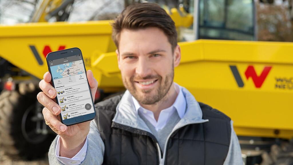 Lächelnder Wacker Neuson Mitarbeiter zeigt Smartphone mit EquipCare Bildschirm.