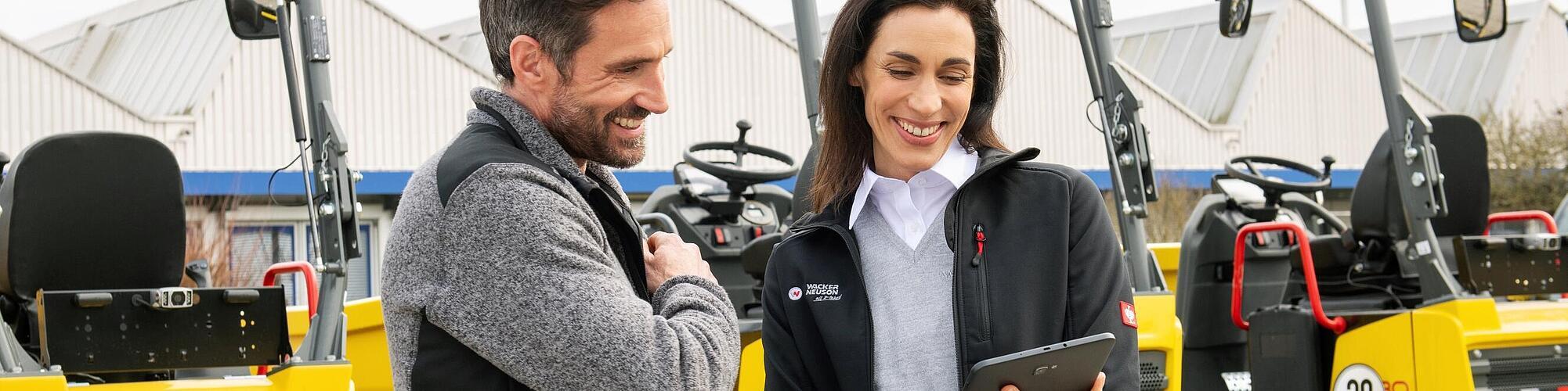 Una sonriente empleada de Wacker Neuson con una tableta asesora a un cliente sonriente frente a las máquinas para la construcción de Wacker Neuson.