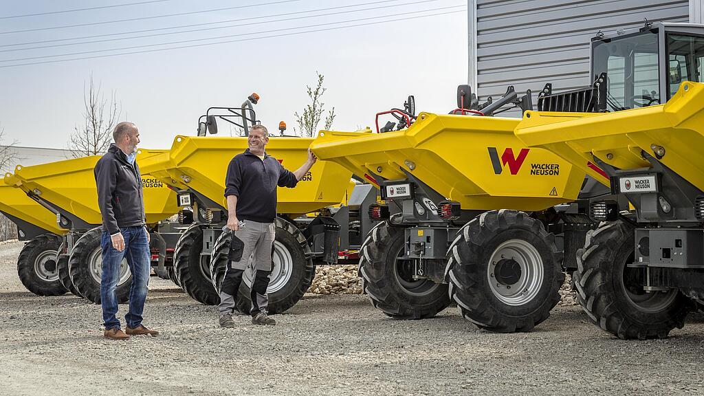 Two men are standing next to several yellow Wacker Neuson dumpers outdoors.