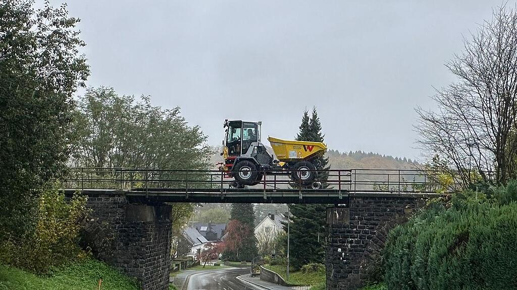 Gelber Wacker Neuson Zweiwege-Baustellen-Dumper fährt über Eisenbahnbrücke über nasse Straße im herbstlichen Dorf