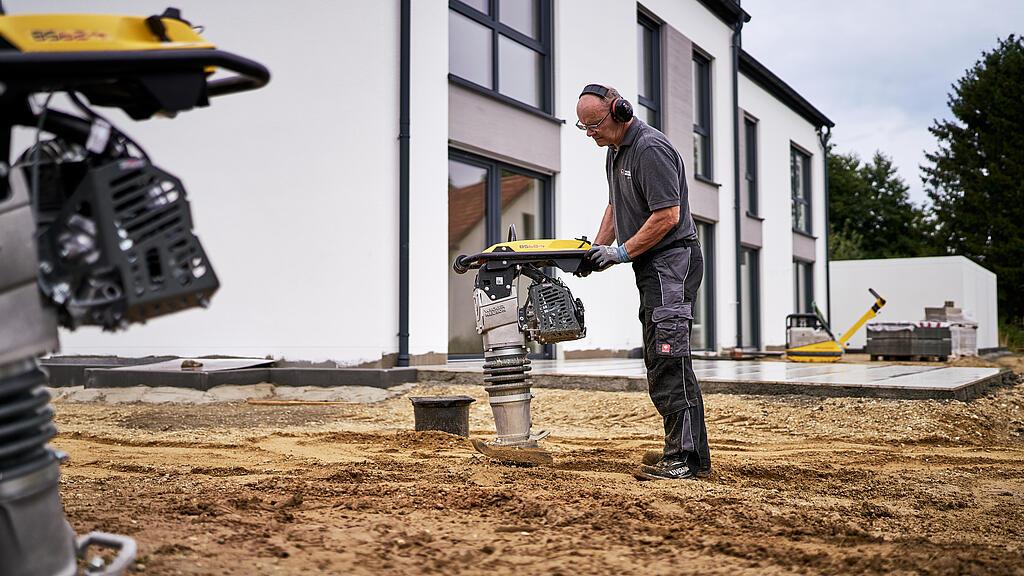 Pracownik zagęszcza grunt przed budynkiem mieszkalnym za pomocą ubijaka wibracyjnego.