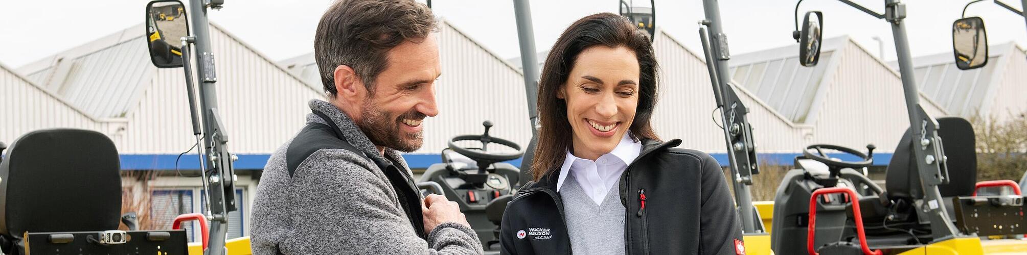 Smiling Wacker Neuson employee with tablet advising smiling customer in front of Wacker Neuson construction machines.