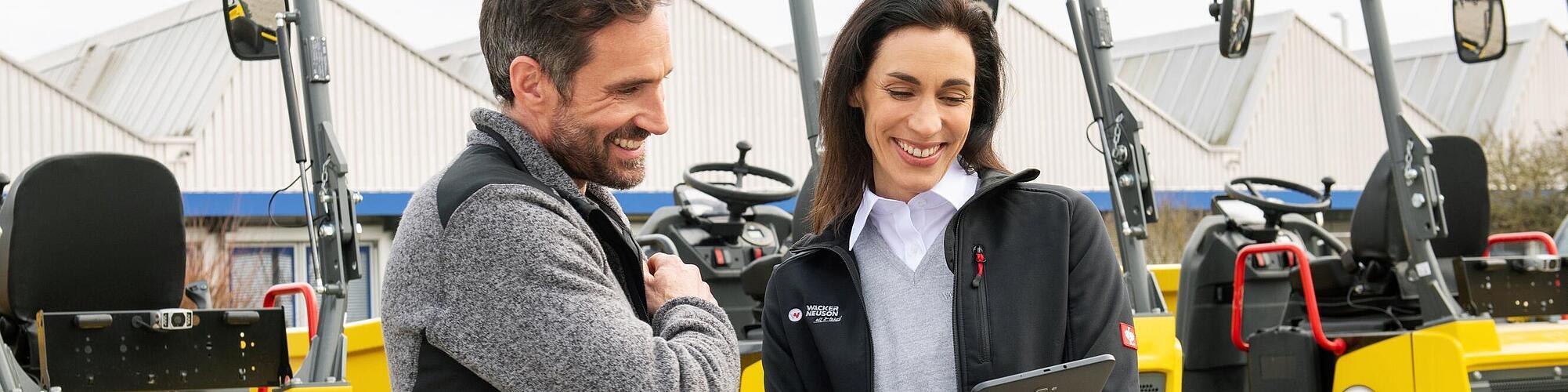 Smiling Wacker Neuson employee with tablet advising smiling customer in front of Wacker Neuson construction machines.