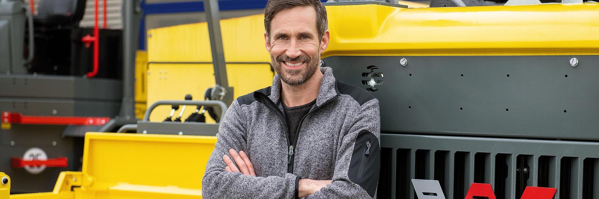 Un empleado sonriente de Wacker Neuson se apoya en una máquina para la construcción de Wacker Neuson con los brazos cruzados.