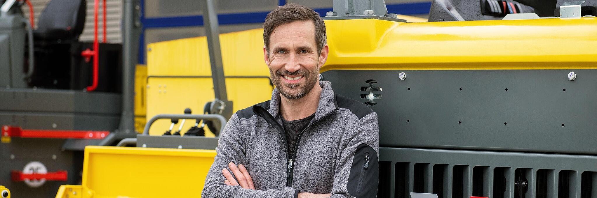 Egy mosolygó Wacker Neuson-munkatárs összefont karral egy Wacker Neuson építőipari gépnek támaszkodik.